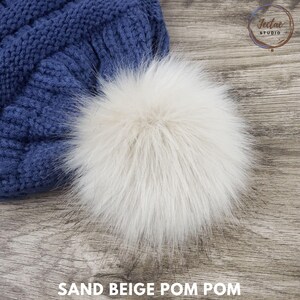 May include: Close-up of a blue knit hat with a large, sand beige pom pom. The pom pom is fluffy and textured. The text "SAND BEIGE POM POM" is at the bottom of the image. The hat is on a wooden surface.