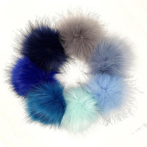 May include: A collection of seven faux fur pom poms in various shades of blue, gray, and light green. The pom poms are arranged in a circle on a white background.