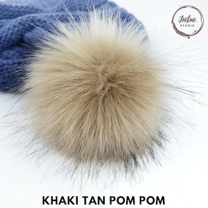 May include: A close-up of a blue knit hat with a large khaki tan pom pom. The pom pom is centered and fluffy, with a mix of tan and dark brown fibers. The text "KHAKI TAN POM POM" is at the bottom.