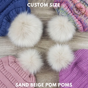 May include: Four sand beige pom poms are displayed on a wooden surface, with the text "CUSTOM SIZE" at the top. The words "SAND BEIGE POM POMS" are at the bottom. The pom poms are fluffy and round, and are next to knitted hats.