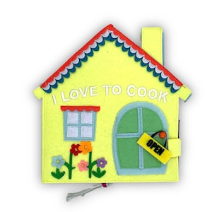 May include: A yellow felt house with a red and blue roof, a green door with a yellow "OPEN" sign, and a window with a white frame. The house has the words "I LOVE TO COOK" on the front. There are green stems with colorful flowers on the front of the house.