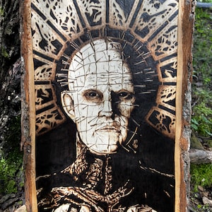Puede incluir: Una rodaja de madera rectangular con un retrato detallado en pirograbado de una figura pálida y calva con numerosos alfileres de metal que sobresalen de la cabeza y la cara, sobre un fondo geométrico oscuro con forma de sol.