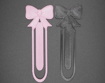 May include: Two 3D printed bookmark designs, one pink and one black, shaped like bows with a long, thin, rectangular base.