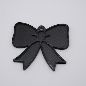 Peut inclure: Ornement noir en forme de nœud papillon avec une surface texturée. Le nœud comporte deux boucles et deux longues queues pointues. L'ornement est probablement en plastique ou dans un matériau similaire.
