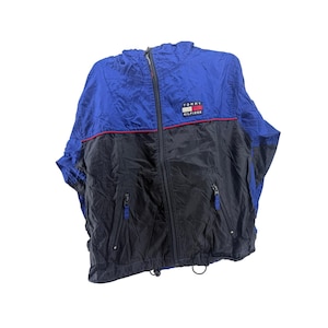 May include: A color-blocked windbreaker jacket featuring a royal blue upper section and a black lower section, separated by a thin red horizontal stripe. The jacket includes a central zipper, a hood, and the Tommy Hilfiger logo on the chest.
