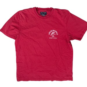 May include: A red short-sleeved t-shirt with a crew neckline. The chest features a white graphic with the text "ABERCROMBIE" arched over an illustration of a chicken, with the text "CHICKEN & FIZZY'S" printed below. An Abercrombie brand label is at the neck.