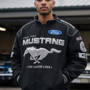 Op de afbeelding: Zwarte Mustang jas met witte accenten, met het Ford logo en een rennend paard embleem. De jas bevat de tekst "SINCE 1964" en "THE LEGEND LIVES". Het heeft een knoopsluiting en zakken.