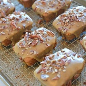 May include: Several mini loaf cakes with a light brown glaze and chopped pecans on top are arranged on a wire rack. The cakes are baked to a golden brown color and are a dessert item.