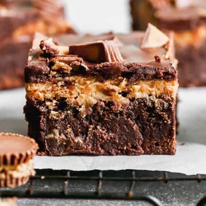 May include: Close-up of a layered dessert featuring a dark chocolate brownie base, a peanut butter filling, and a chocolate topping. The dessert is garnished with peanut butter cups and pieces. The image is shot from a high angle.