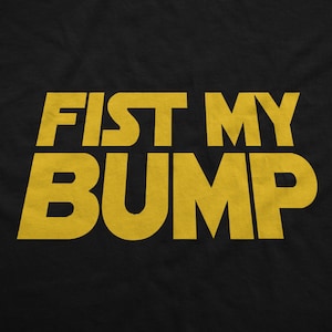 May include: Black t-shirt featuring the phrase "FIST MY BUMP" in large, yellow, block letters. The text is centrally aligned and dominates the image. The font is bold and has a slightly worn appearance.