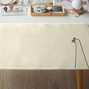Può includere: Un tappetino da scrivania color crema con un semplice design a fiore e uccello. Il tappetino è su una scrivania bianca con una gamba di tavolo in legno.