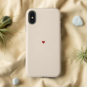 Puede incluir: Funda de teléfono beige claro con un pequeño corazón rojo en el centro. La funda del teléfono tiene una cámara de doble lente. El fondo incluye plantas aéreas y pequeñas piedras decorativas sobre una tela beige.