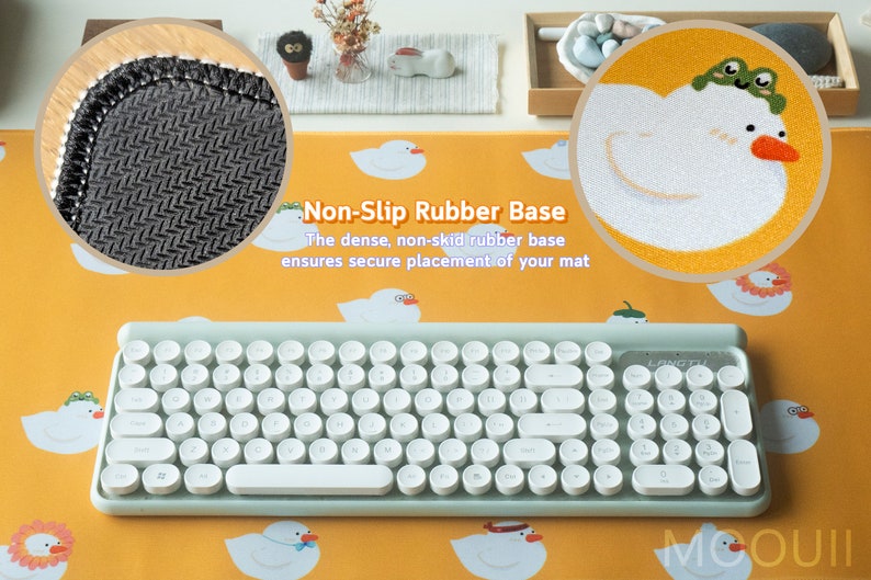 May include: A light blue keyboard with round white keys and a non-slip rubber base. The keyboard is on a yellow desk mat with a white duck design. The text "Non-Slip Rubber Base" is written in black on the mat. The text "The dense, non-skid rubber base ensures secure placement of your mat" is written in black below the title.