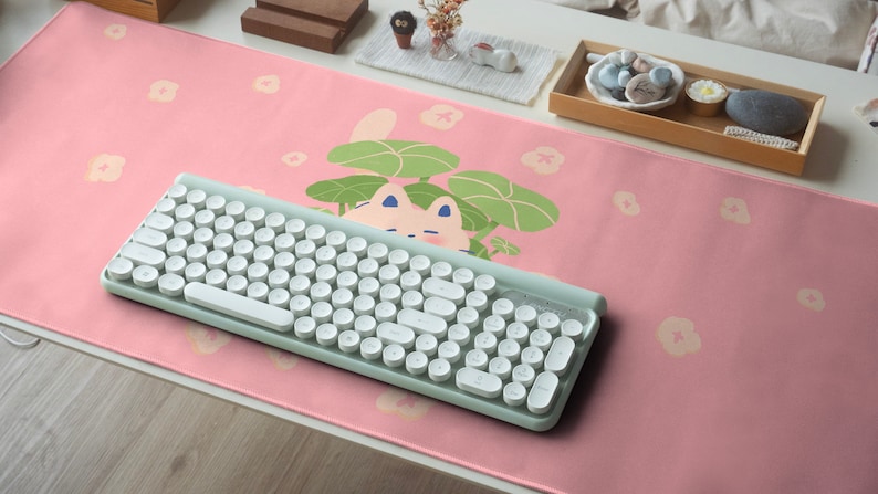 May include: A pink desk mat with a white cat illustration and a light green keyboard. The mat has a floral pattern with white flowers.