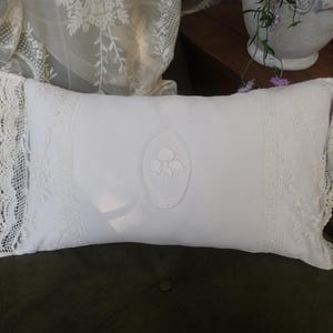 Può includere: Un cuscino rettangolare bianco con un'intricata bordatura in pizzo e un motivo floreale ricamato al centro. Il cuscino è appoggiato su una superficie verde scuro, con uno sfondo di tessuto di pizzo e mobili in legno.
