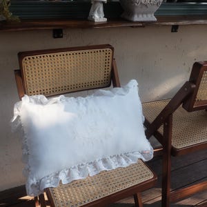 Puede incluir: Un cojín rectangular blanco con un diseño floral y ribete de encaje descansa sobre una silla de madera con asiento de mimbre tejido. La silla es marrón y tiene un diseño plegable. El cojín es el punto focal.