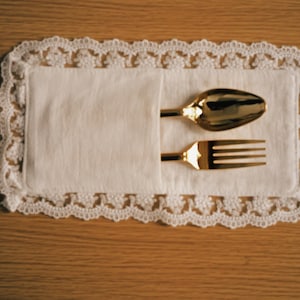 Könnte beinhalten: Ein goldener Löffel und eine Gabel liegen auf einer weißen Leinen-Serviette, eingebettet in eine dekorative, weiß spitzenbesetzte Tischdecke. Das Besteck glänzt vor dem neutralen Hintergrund der Serviette und der Holzoberfläche.