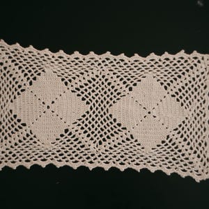 May include: Cream-coloured, rectangular doily with a crocheted openwork design. The doily features two central, solid, diamond-shaped patterns and a scalloped edge. The intricate lace-like pattern is visible against a dark background.