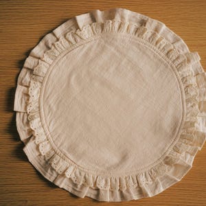 Op de afbeelding: Een ronde, crèmekleurige placemat met een gerimpelde rand. De placemat heeft een eenvoudig, elegant ontwerp, met een effen midden en een decoratieve rand. De gerimpelde rand voegt textuur en visuele interesse toe. De placemat is gemaakt van een zachte, natuurlijk ogende stof.