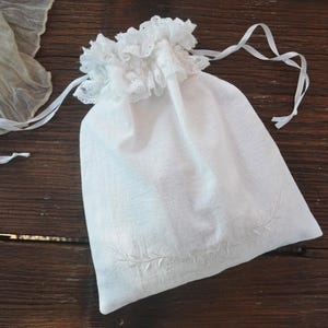 May include: A white linen drawstring bag with a ruffled lace trim at the top. The bag has a delicate embroidered leaf design near the bottom. White satin ribbons are attached for closure. The bag is on a dark wood surface.