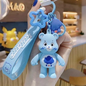 May include: A light blue Care Bear keychain featuring a grumpy bear with a cloud belly badge. The keychain includes a star-shaped charm and a strap with "Care Bears" text and a bear graphic. The bear is approximately 2 inches tall.