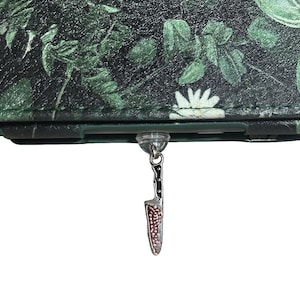 May include: A dark green wallet with a botanical leaf pattern. A small knife charm with a red and silver blade hangs from the wallet. The wallet has a clear plastic attachment.