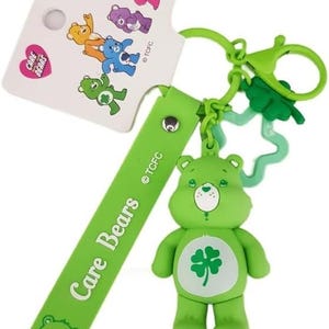 Puede incluir: Un llavero verde de Care Bears con un trébol de cuatro hojas en el vientre. El llavero incluye una correa verde con "Care Bears" impreso, un clip verde y una etiqueta blanca con personajes de Care Bears.