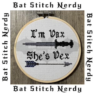 Op de afbeelding: Een witte borduurring met een kruissteekontwerp met de tekst "I'm Vax She's Vex". Het ontwerp bevat een zwart dolk en een blauwe en zwarte pijl.