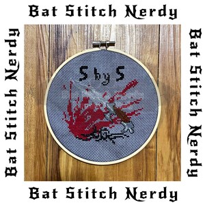 May include: A close-up of a cross-stitch embroidery hoop with a red and black design. The hoop is on a wooden surface. The embroidery is titled "5 by 5".