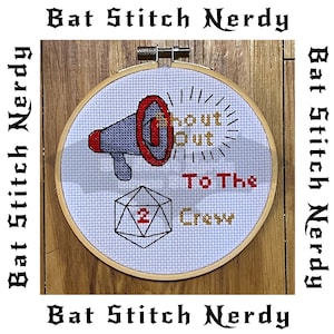 May include: A white cross-stitch embroidery hoop with a red and grey megaphone, a gold 20-sided die, and the text "Shout Out To The Crew" in gold and red thread.