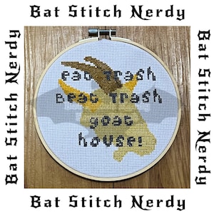 May include: A white embroidery hoop with a cross-stitch design of a goat with the text "Eat Trash Beat Trash Goat House!" in yellow and brown thread.