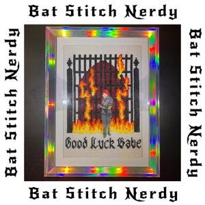 Puede incluir: Obra de arte de punto de cruz enmarcada con una persona frente a una puerta en llamas. La obra presenta el texto "Good Luck Babe" y está enmarcada en un borde holográfico. Las palabras "Bat Stitch Nerdy" están impresas alrededor del marco.