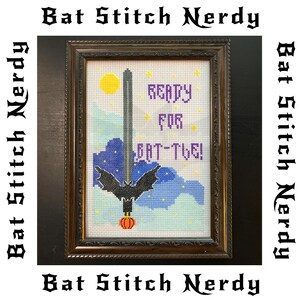 May include: A framed cross-stitch design with a purple bat and the text "READY FOR BAT-TIVE!" in a purple font. The bat is hanging from a sword-like object with a yellow moon in the background.