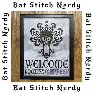 May include: A framed cross-stitch design featuring a black and white silhouette of a monster with the text "WELCOME FOOLISH MUPPETS" in black and purple lettering.