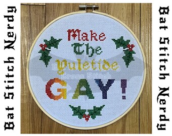 May include: A white embroidery hoop with a cross-stitch design that reads "Make the Yuletide GAY!" in a rainbow font. The design is surrounded by green holly leaves with red berries.