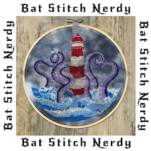 May include: A cross-stitch embroidery hoop with a lighthouse and an octopus. The lighthouse is red and white striped and the octopus is purple. The background is blue and white to represent the ocean.