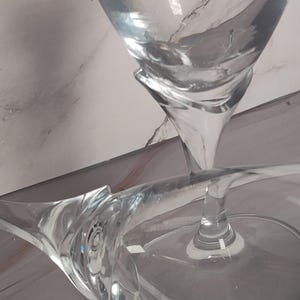 May include: Clear glass stemware with a unique, curved design. The glasses have a wide bowl and a slender stem, perfect for serving beverages. The image shows the glasses at an angle, highlighting their elegant shape.