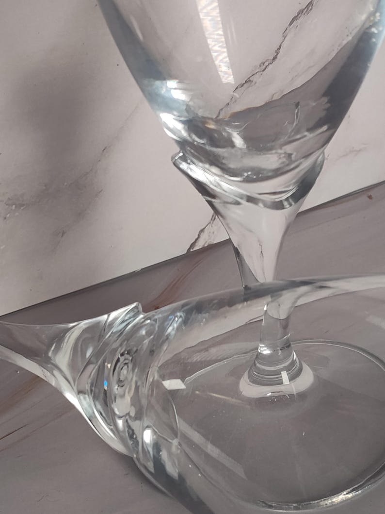 May include: Clear glass stemware with a unique, curved design. The glasses have a wide bowl and a slender stem, perfect for serving beverages. The image shows the glasses at an angle, highlighting their elegant shape.