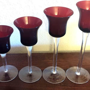 May include: Four elegant, stemmed candle holders. Each has a clear, slender stem and a circular base. The bowls are a deep burgundy color, with a flared, tulip-like shape. The candle holders are arranged in a row.