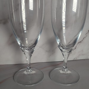 May include: Two clear, stemmed wine glasses. The glasses have a rounded bowl shape and a slender stem that connects to a circular base. The glasses are made of transparent glass, reflecting light. The background is a light gray and white marble pattern.