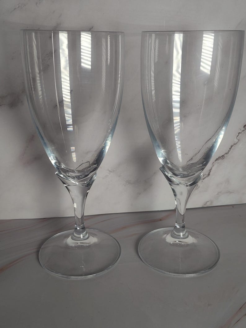 May include: Two clear, stemmed wine glasses. The glasses have a rounded bowl shape and a slender stem that connects to a circular base. The glasses are made of transparent glass, reflecting light. The background is a light gray and white marble pattern.