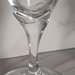 May include: A clear, stemmed martini glass with a wide, triangular bowl and a slender stem. The glass has a unique design element where the bowl meets the stem, with a swirling, decorative feature. The base is round and flat.