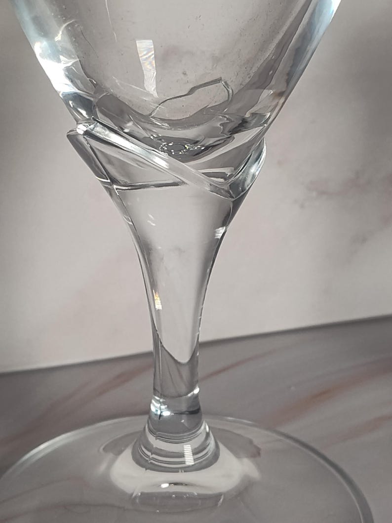 May include: A clear, stemmed martini glass with a wide, triangular bowl and a slender stem. The glass has a unique design element where the bowl meets the stem, with a swirling, decorative feature. The base is round and flat.