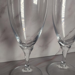 May include: Two clear champagne flutes with a twisted stem design. The glasses have a wide, circular base and a tall, slender bowl. The background is a light-colored surface with subtle veining, and the glasses reflect light.