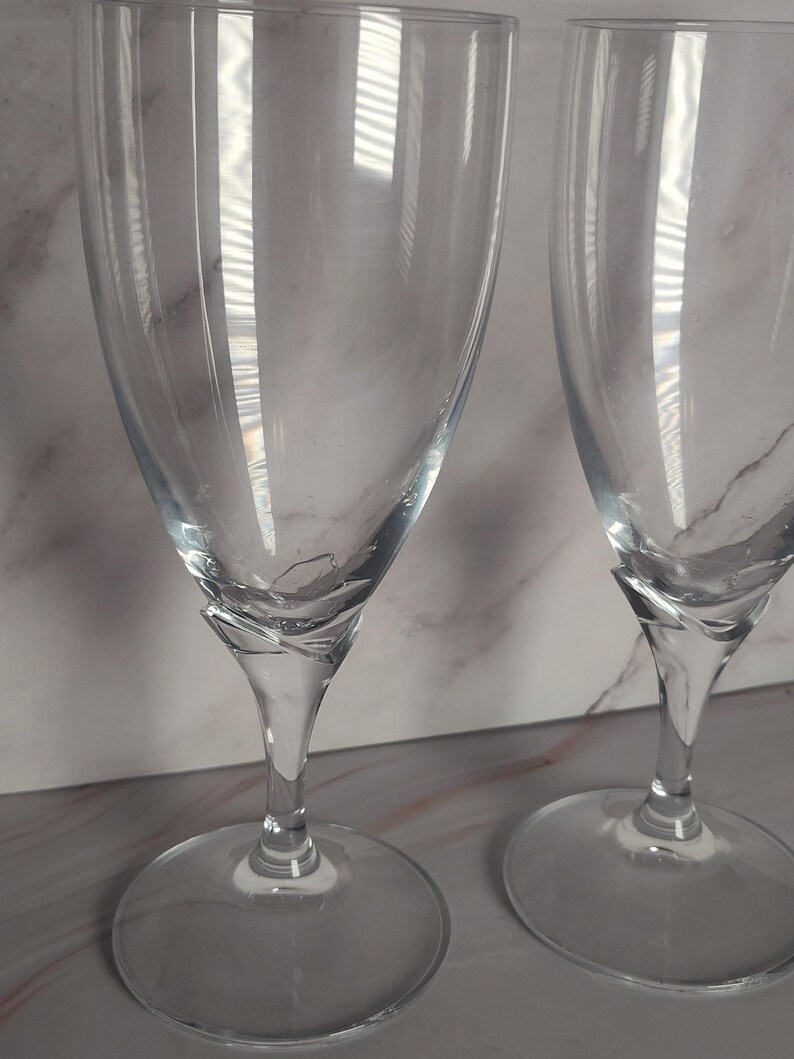 May include: Two clear champagne flutes with a twisted stem design. The glasses have a wide, circular base and a tall, slender bowl. The background is a light-colored surface with subtle veining, and the glasses reflect light.