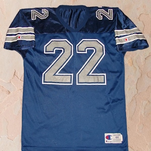 Puede incluir: Una camiseta de f&uacute;tbol azul marino con n&uacute;meros plateados "22" en la parte delantera y "2" en los hombros. La camiseta tiene rayas plateadas y blancas en las mangas y un logotipo de Champion en la manga derecha y en la esquina inferior derecha.