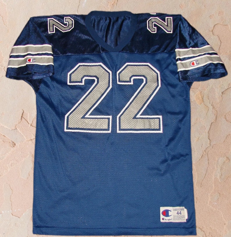 Puede incluir: Una camiseta de f&uacute;tbol azul marino con n&uacute;meros plateados "22" en la parte delantera y "2" en los hombros. La camiseta tiene rayas plateadas y blancas en las mangas y un logotipo de Champion en la manga derecha y en la esquina inferior derecha.