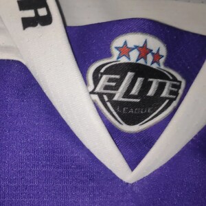 May include: Purple and white hockey jersey with a black patch on the front that says "Elite League" with three stars above the text.