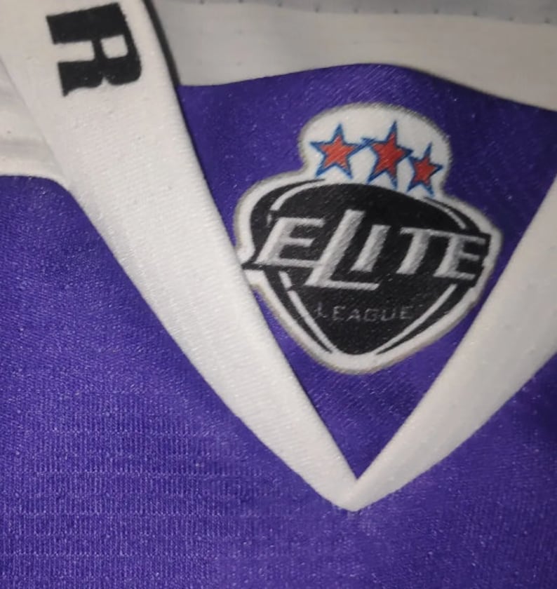 May include: Purple and white hockey jersey with a black patch on the front that says "Elite League" with three stars above the text.