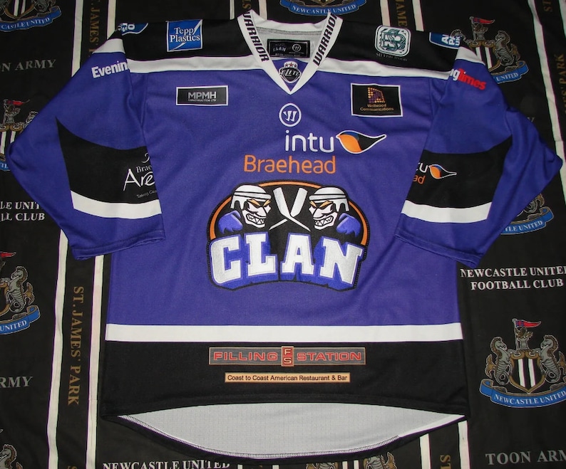 May include: Purple hockey jersey with black and white trim. The front of the jersey features a large logo with two hockey players wearing helmets and the word "CLAN" in blue. The jersey also has the words "intu Braehead" and "Filling Station" on the front.